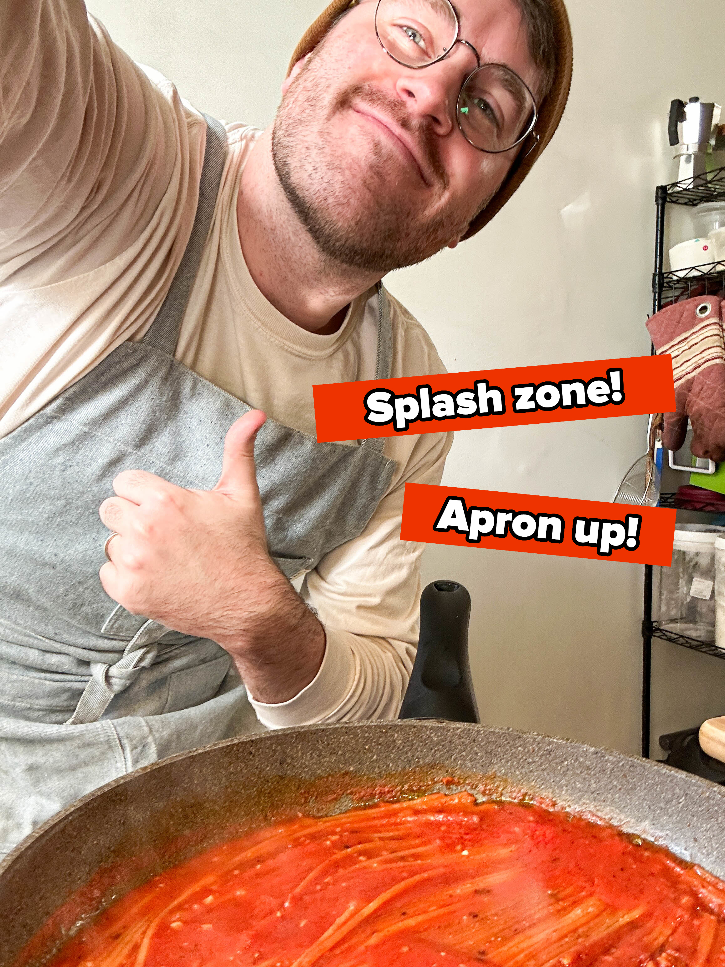 Person giving a thumbs-up over a large pan of sauce. They wear an apron and glasses, smiling at the camera with text saying splash zone and apron up