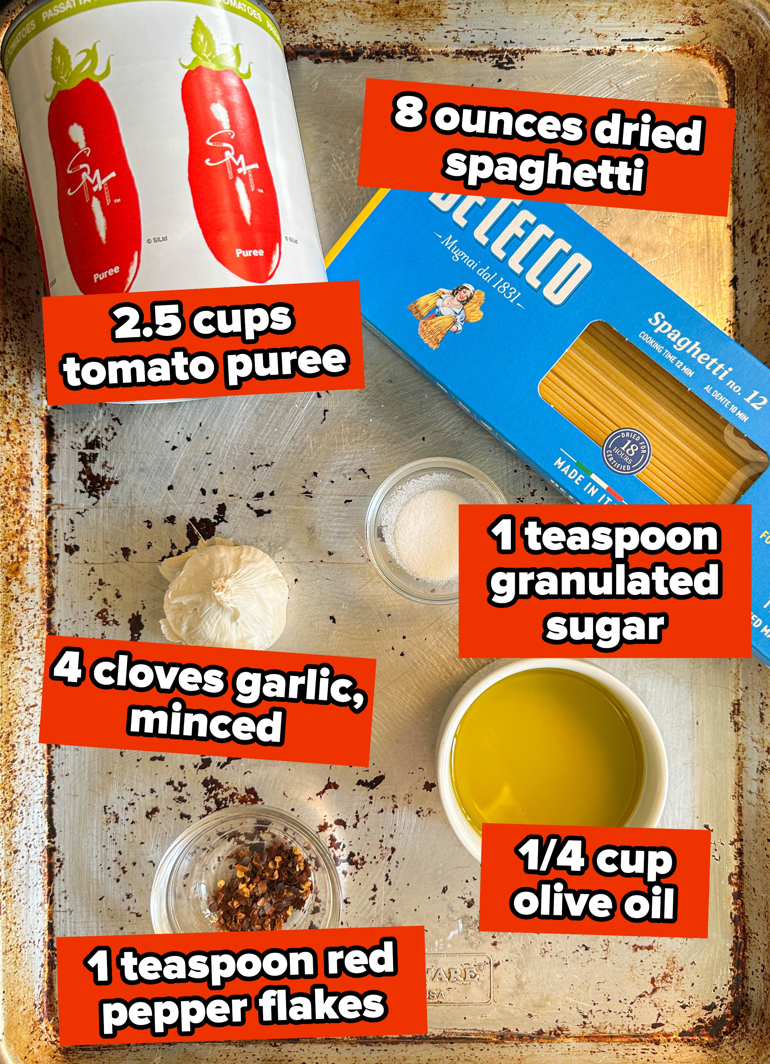 Ingredients for a recipe laid out on a baking sheet including canned tomatoes, pasta, garlic, oil, sugar, and pepper flakes