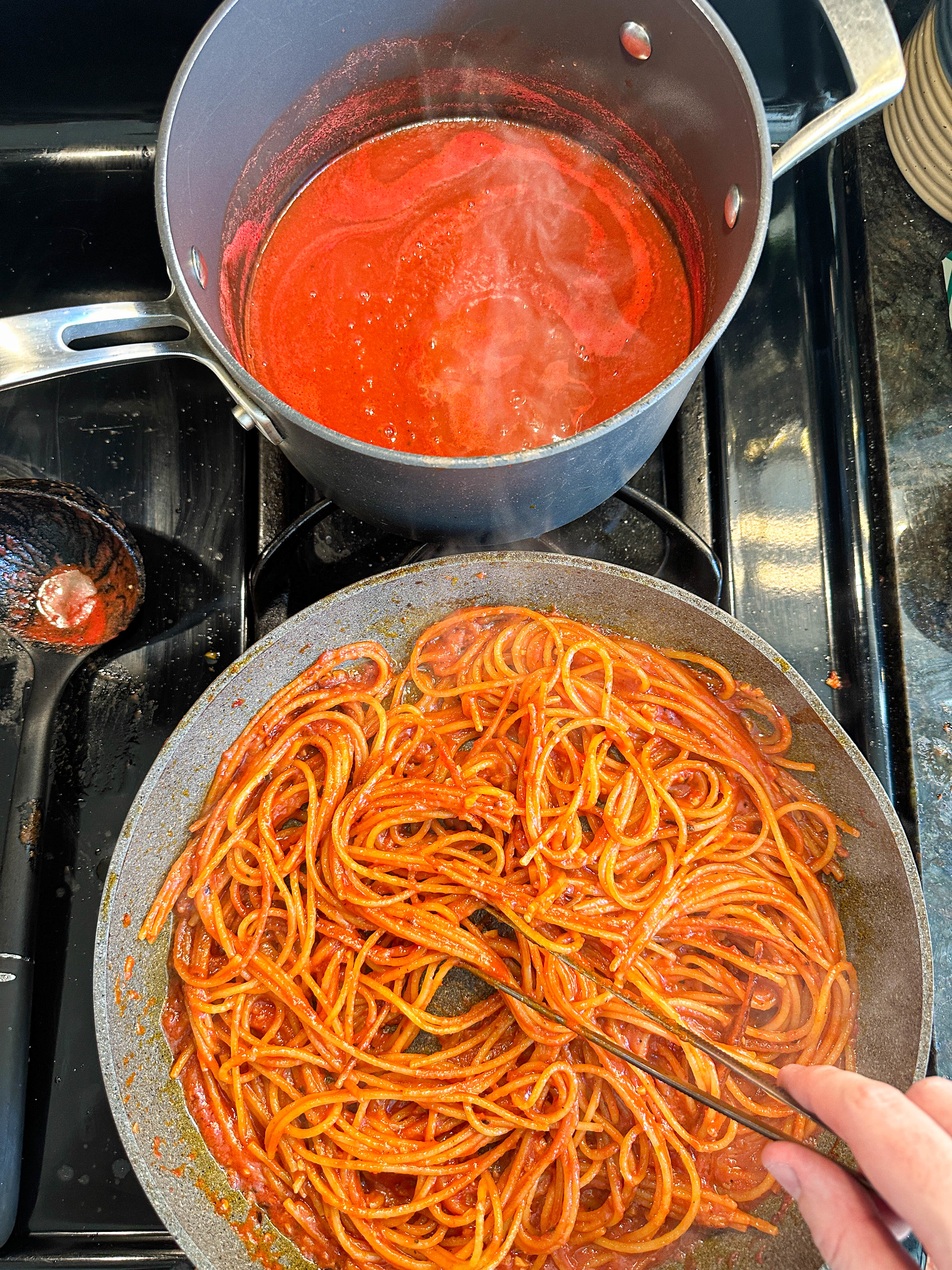 A person stirring spaghetti with tomato sauce in a pan, next to a pot of sauce boiling over on a stovetop