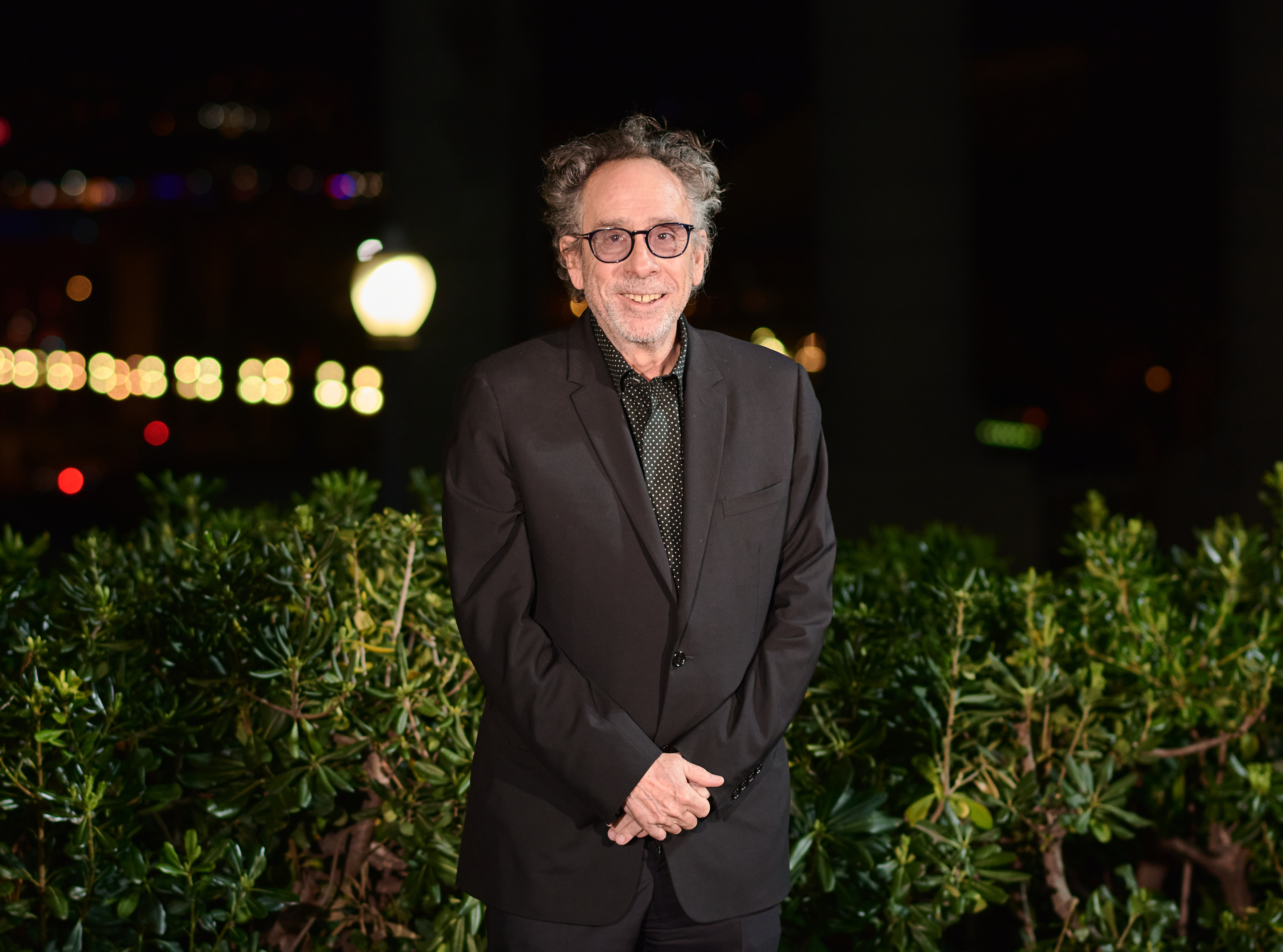 Tim Burton standing before foliage, wearing a suit with a patterned shirt and no tie