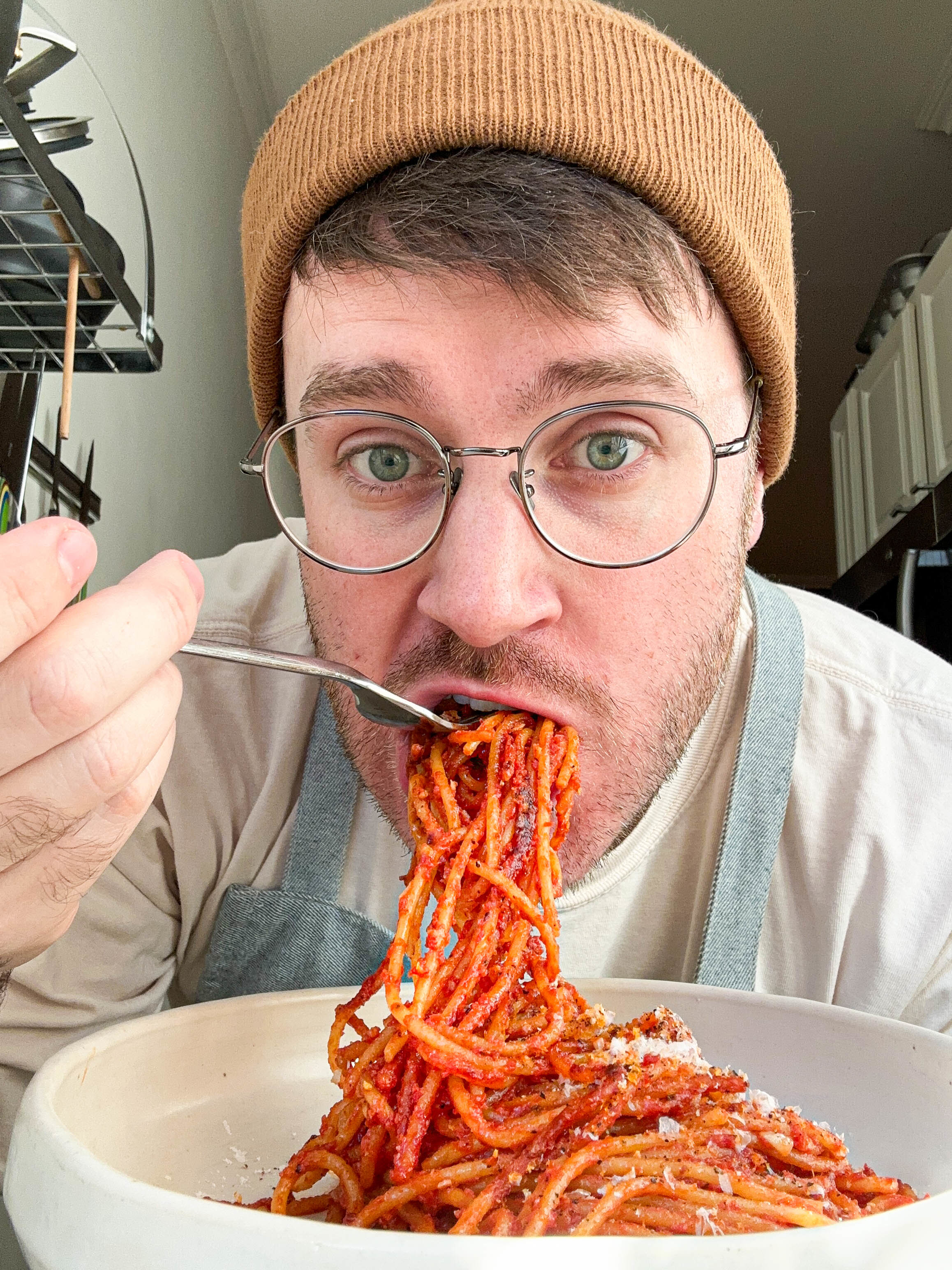 Man with glasses and beanie eating spaghetti, looking at camera