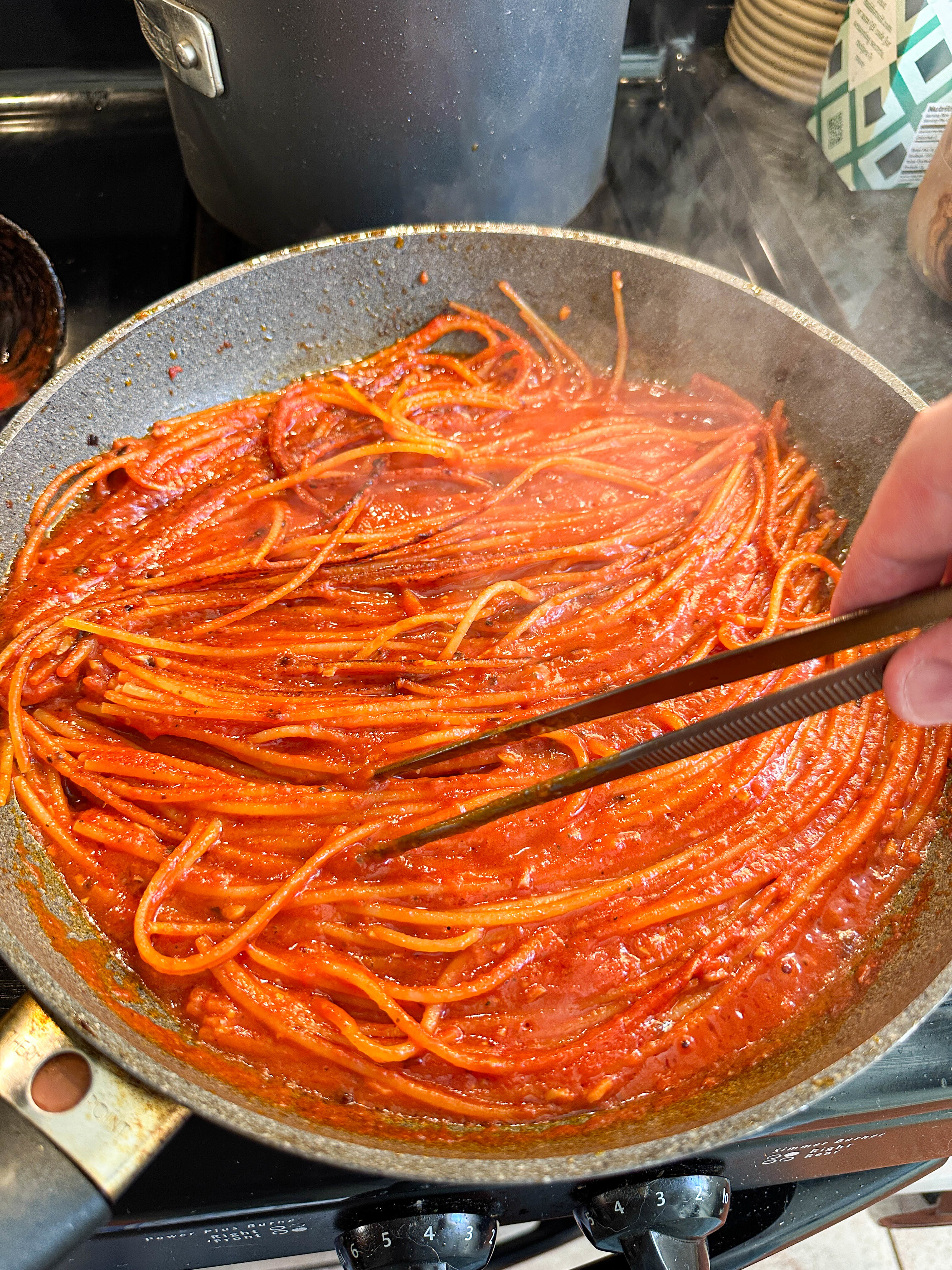 A person cooking spaghetti in tomato sauce in a pan, using tongs to stir