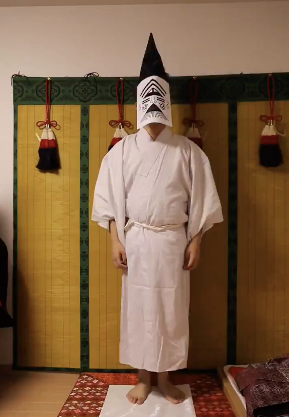 和室にいる人物が白い着物を着て、顔には伝統的な紙のお面をかぶっています。