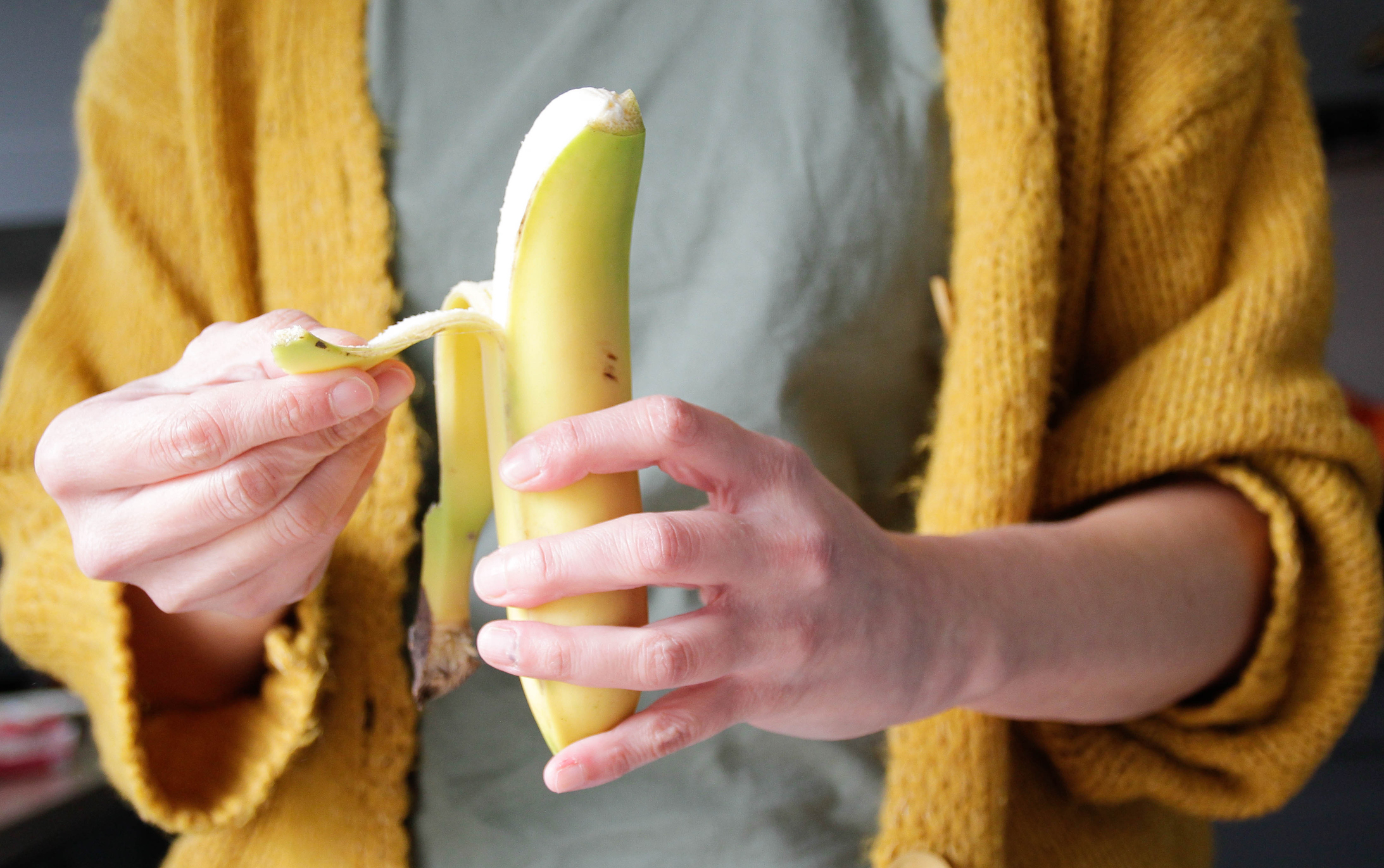 Person peeling a banana, wearing a yellow cardigan over a grey shirt