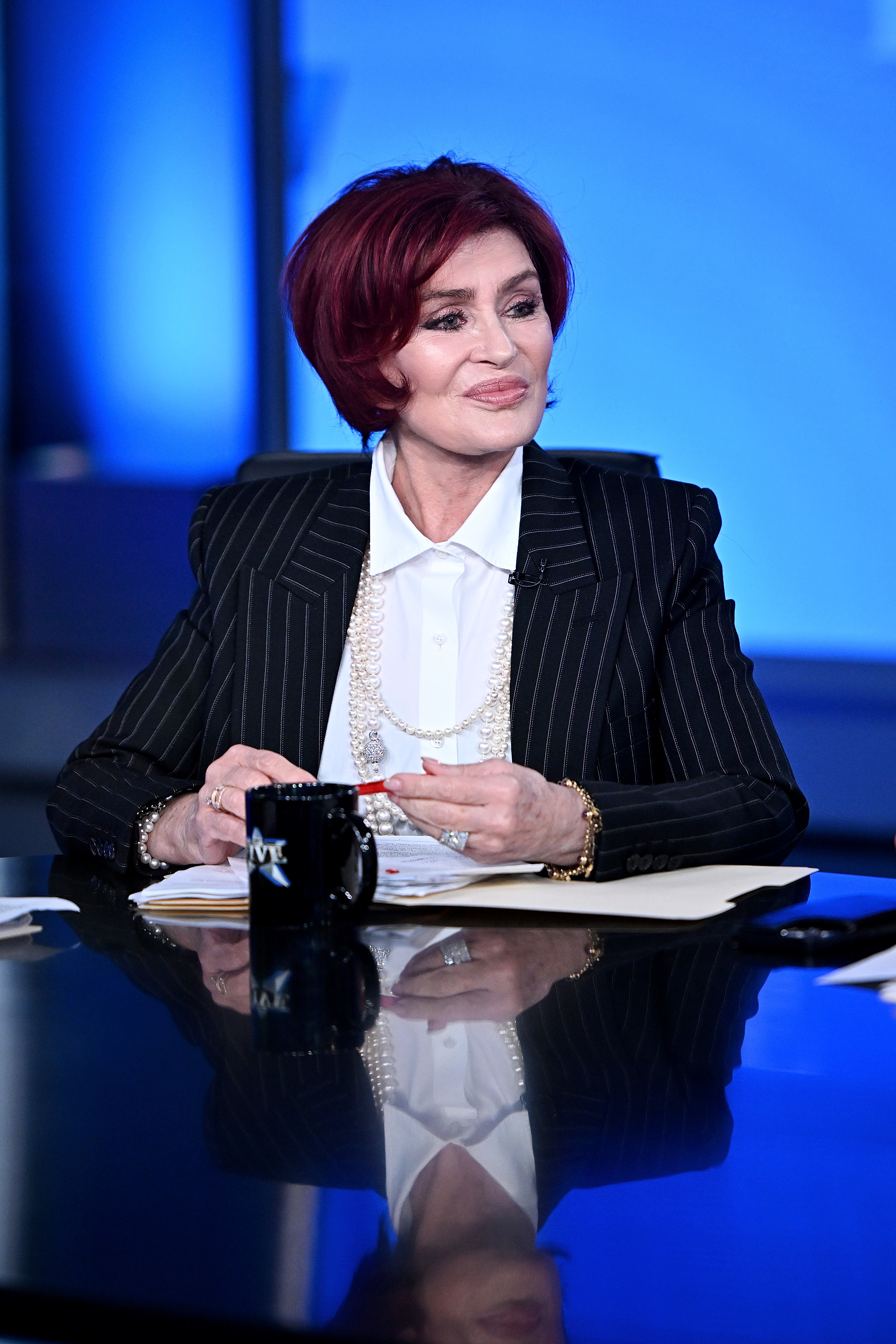 Sharon Osbourne seated at a desk wearing a pin-striped suit and layered necklaces