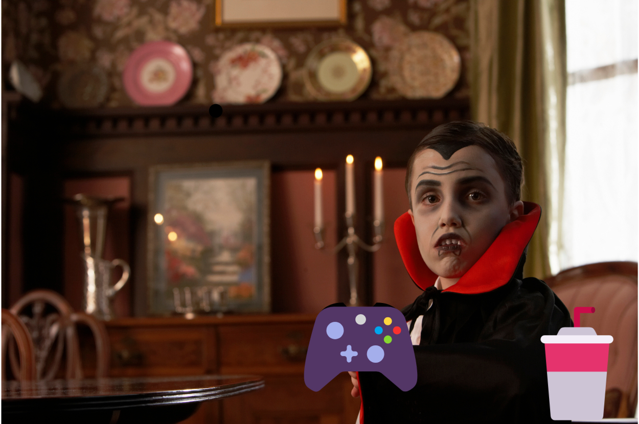 Person in vampire costume sits at a table with a game controller, with a drink to the right