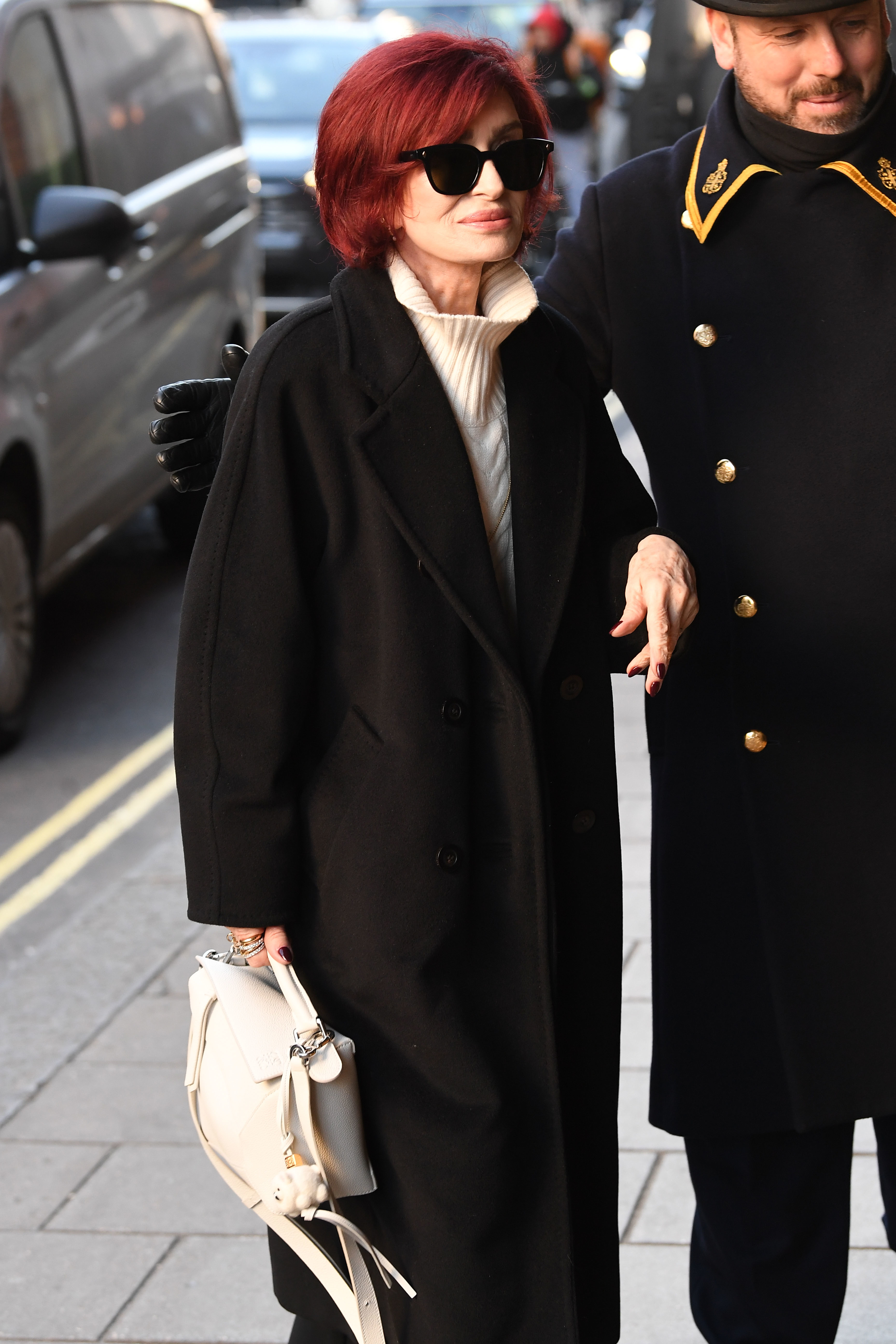 Sharon in a coat with a hite handbag escorted by a man into a building