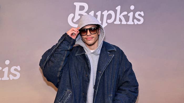 Person in a denim jacket and beanie poses with sunglasses at the &#x27;Bupkis&#x27; event backdrop