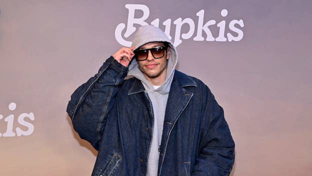 Person in a denim jacket and beanie poses with sunglasses at the 'Bupkis' event backdrop