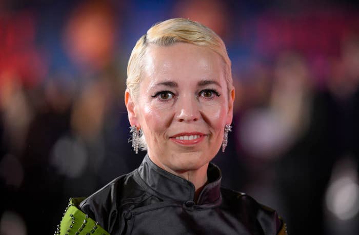 Close-up of Olivia at a gala, smiling, with a unique updo hairstyle and jeweled earrings, wearing a high-collar black outfit