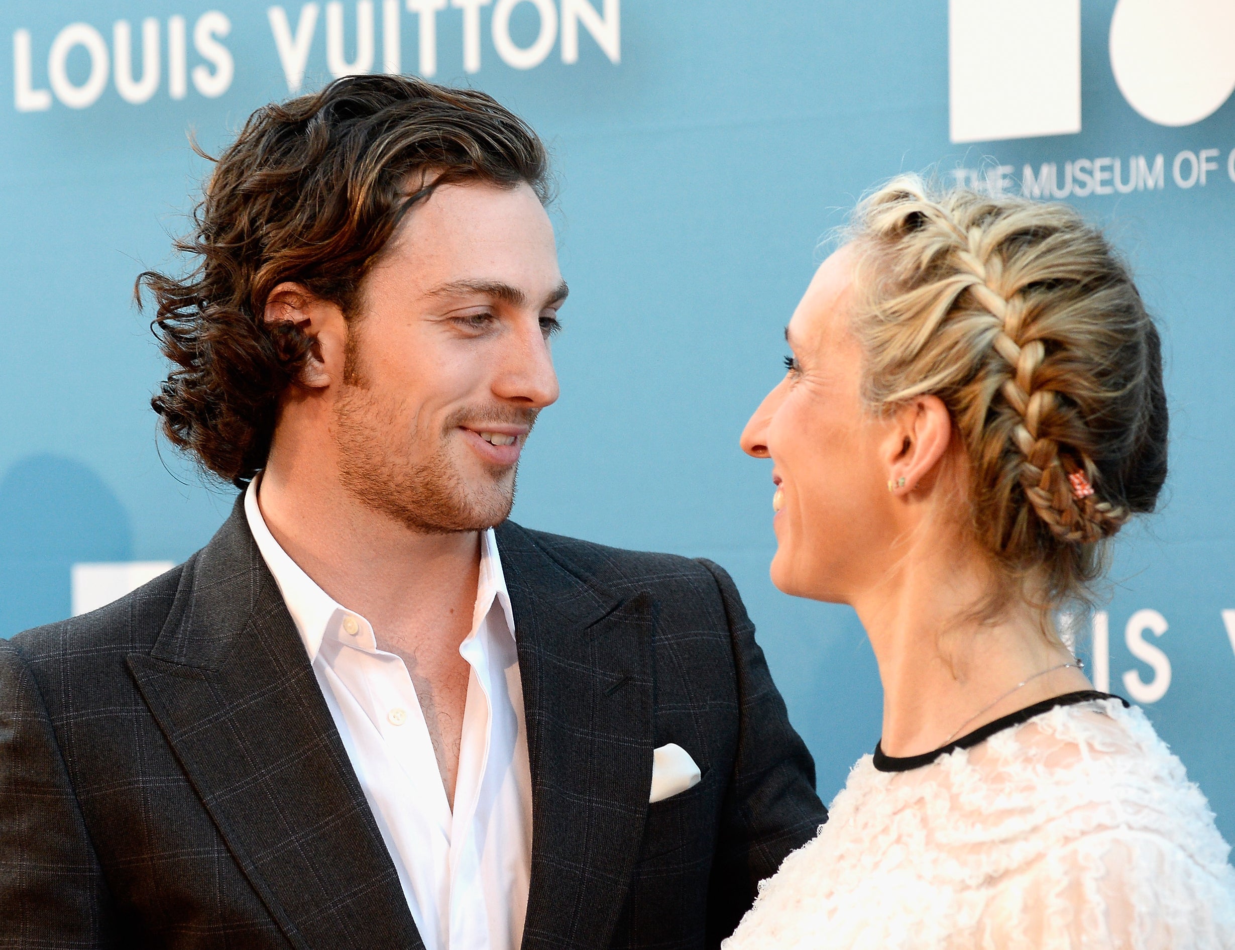 The couple smiling at each other, he in a suit and she in textured dress, at a media event