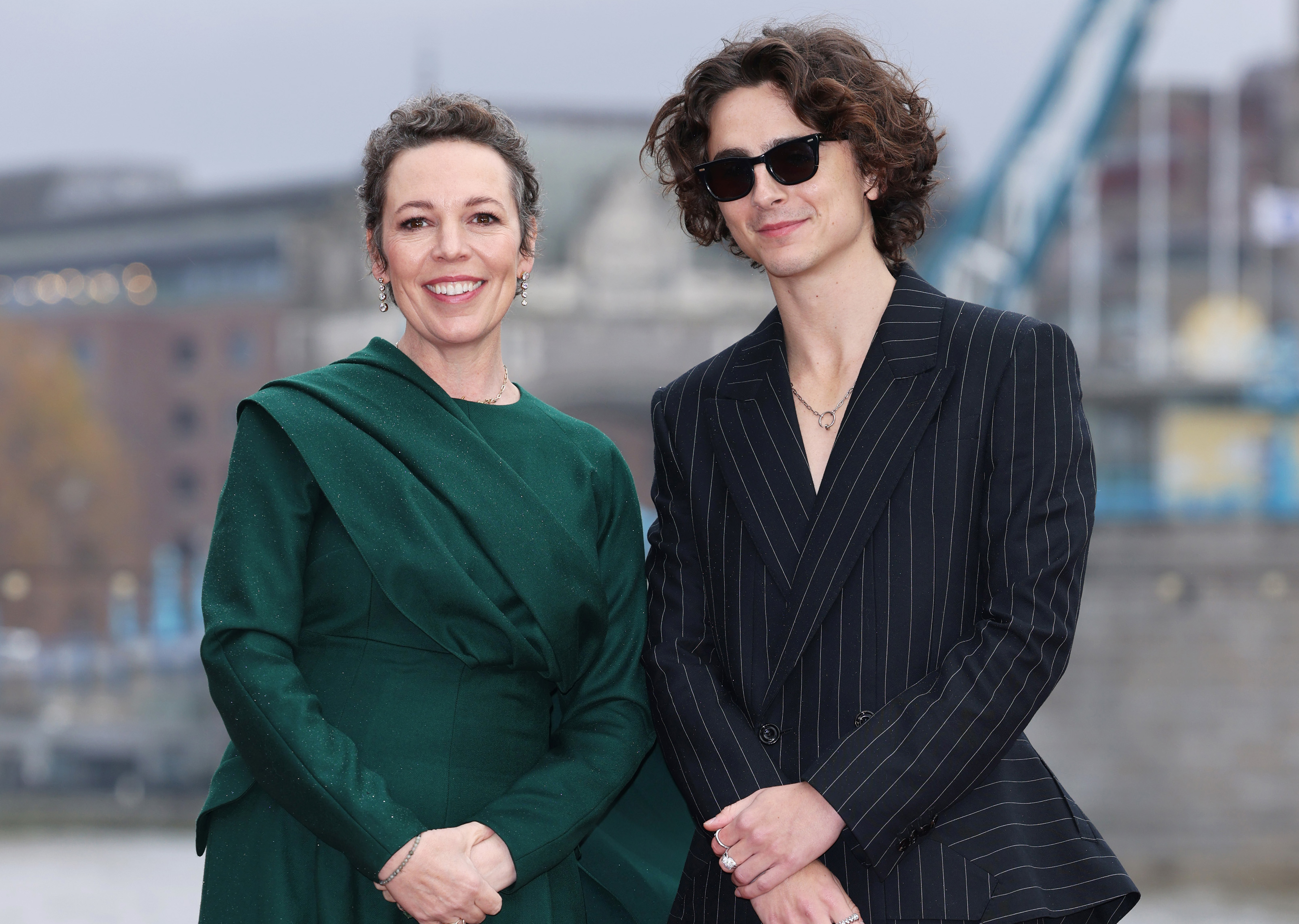 Olivia smiling in a long-sleeved dress with draped detail stands next to Timothée Chalamet, in a pin-striped suit, also smiling