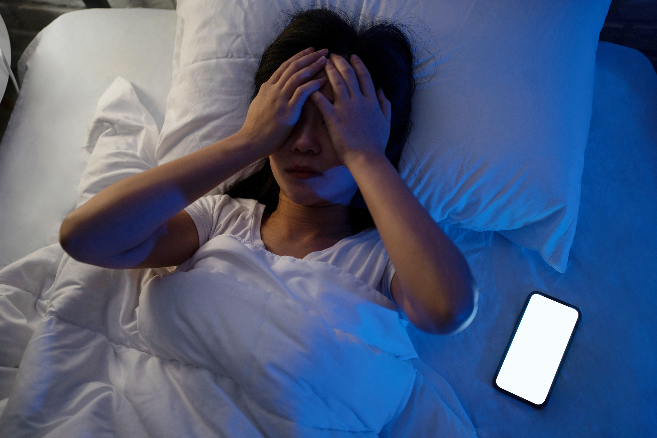 Woman in bed covering face with hands, phone lit on the side, suggesting insomnia or stress