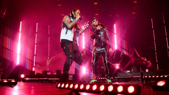 Two musicians performing on stage, one in a vest and the other in a leather jacket, with bright stage lighting in the background