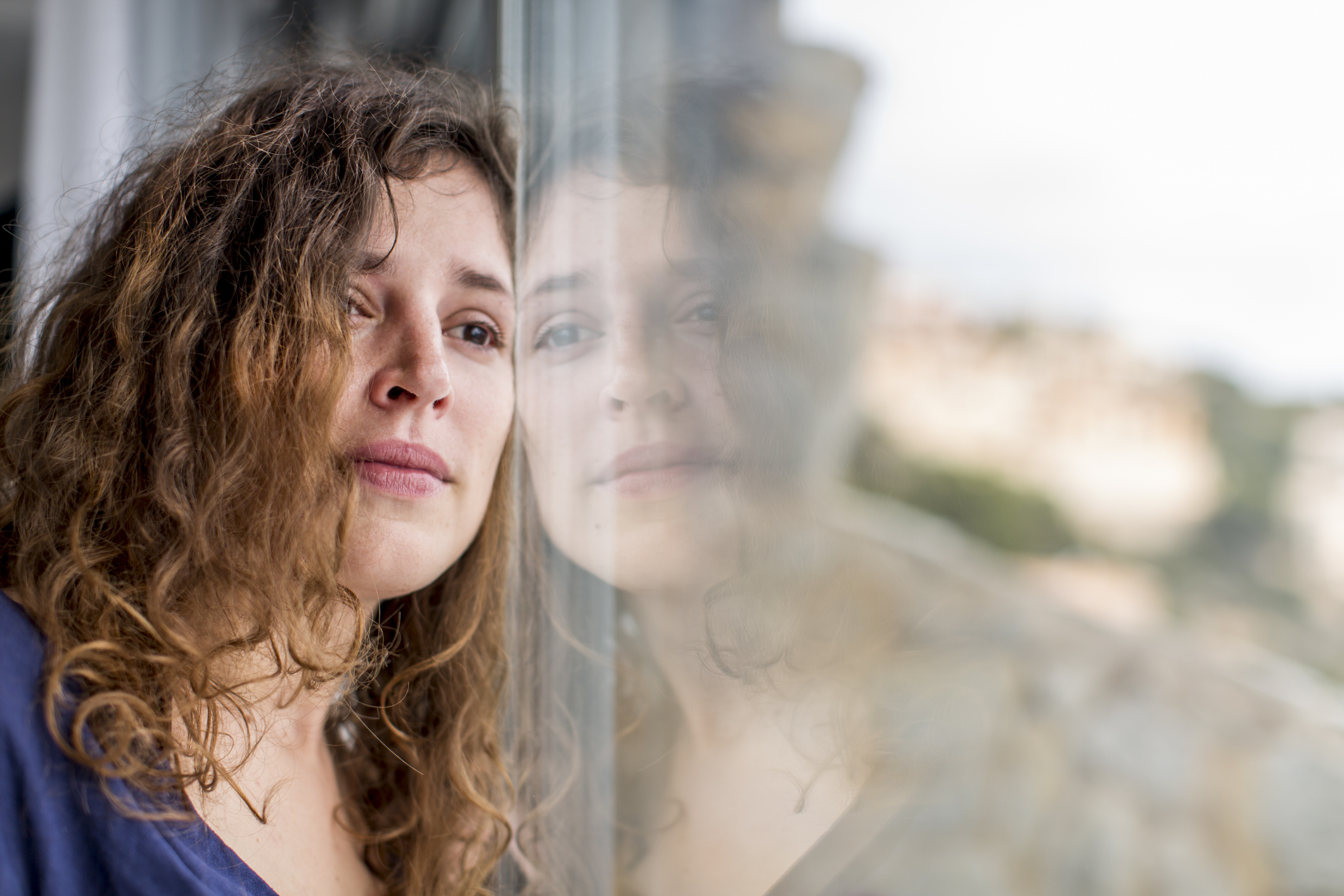 Woman with wavy hair resting her head against a window, reflected on the glass, looking thoughtful