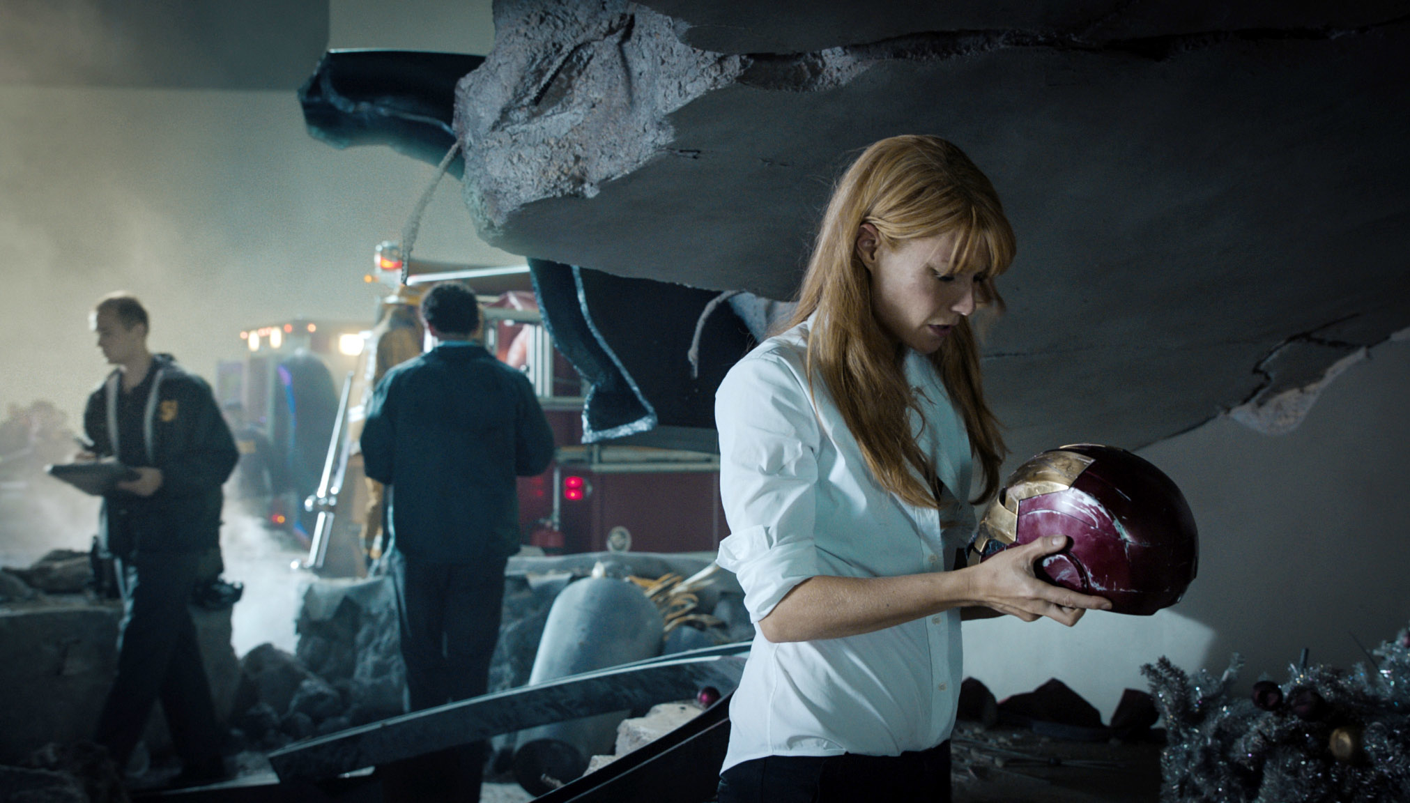 Pepper Potts examining a Iron Man&#x27;s helmet amid debris with emergency personnel in background