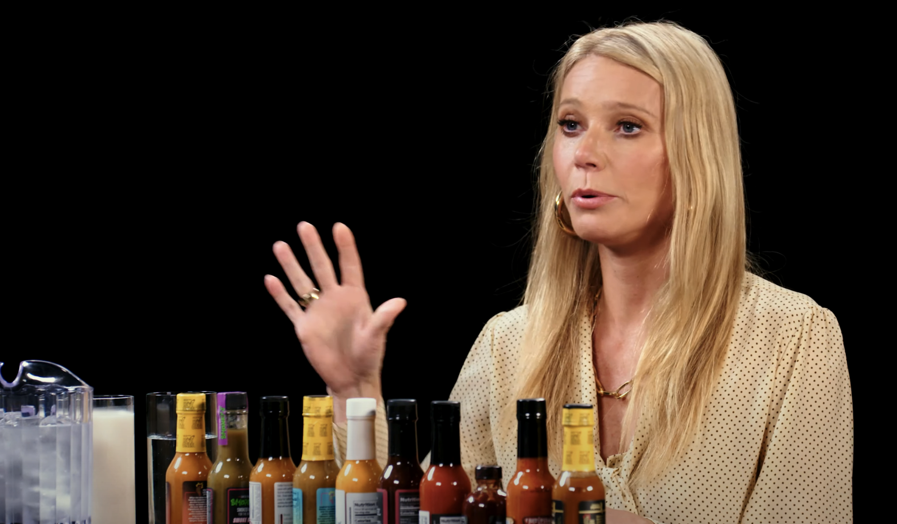 Gwyneth gesturing during an interview, with a lineup of hot sauce bottles on the table