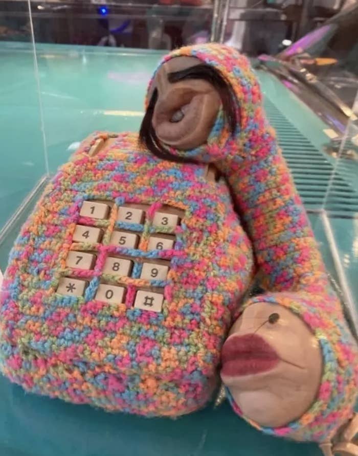 Crocheted phone in the shape of a person, with keypads on the chest and two head-like ends, displayed on glass