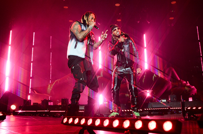 Two musicians performing on stage, one in a vest and the other in a leather jacket, with bright stage lighting in the background
