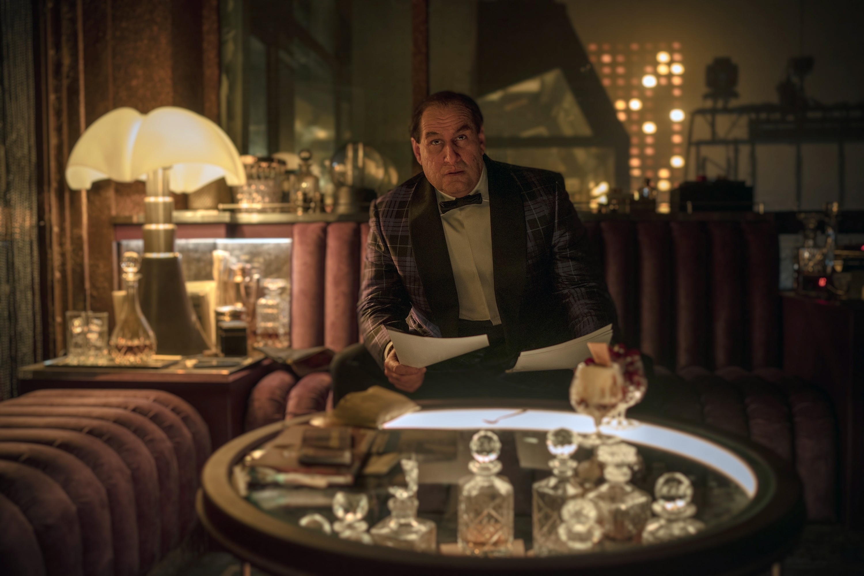 Penguin in a plaid suit sitting at a circular booth with papers in a dimly lit room with a cityscape background