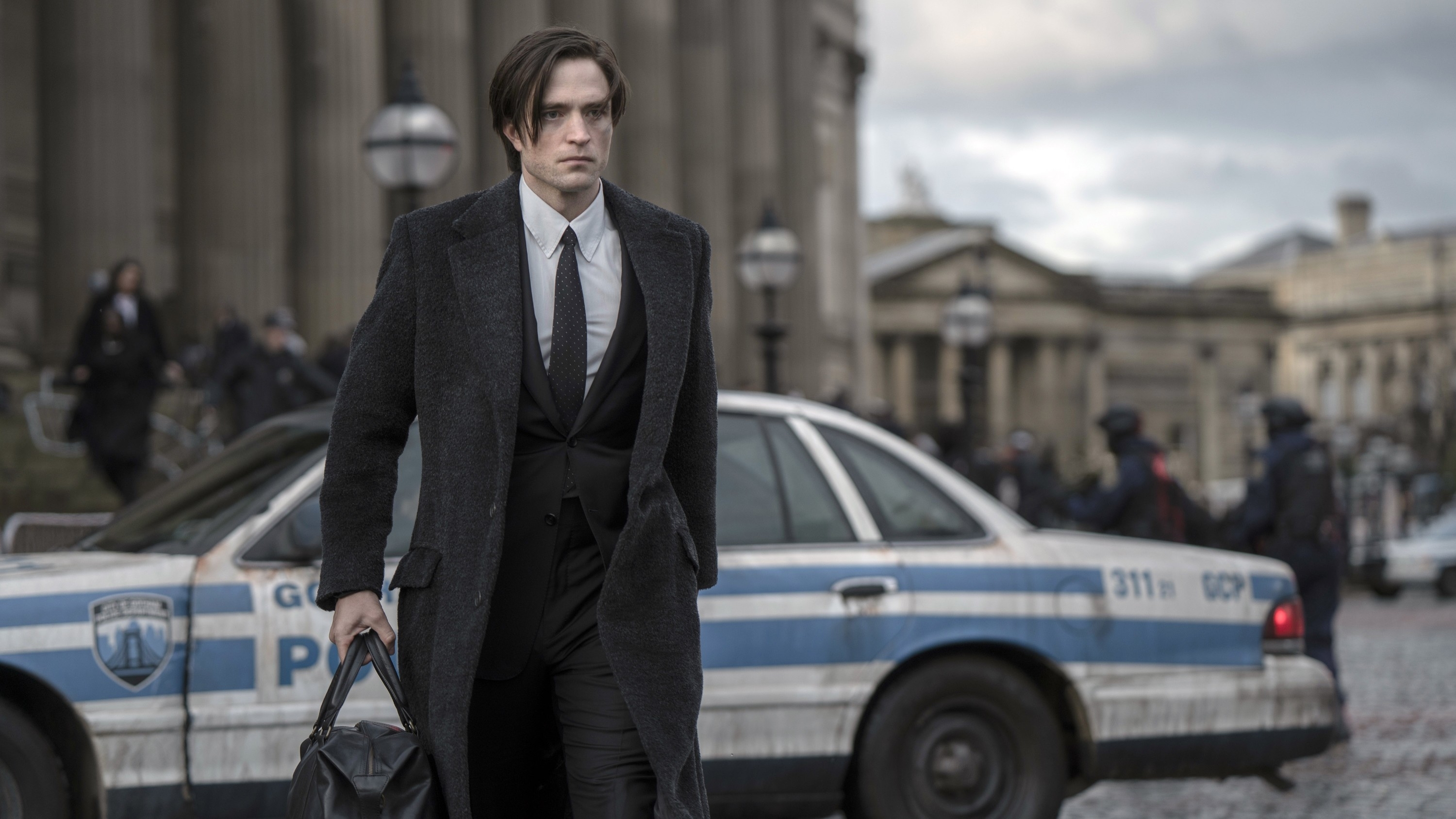 Robert Pattinson in a suit and overcoat walking in front of a police car and classical architecture