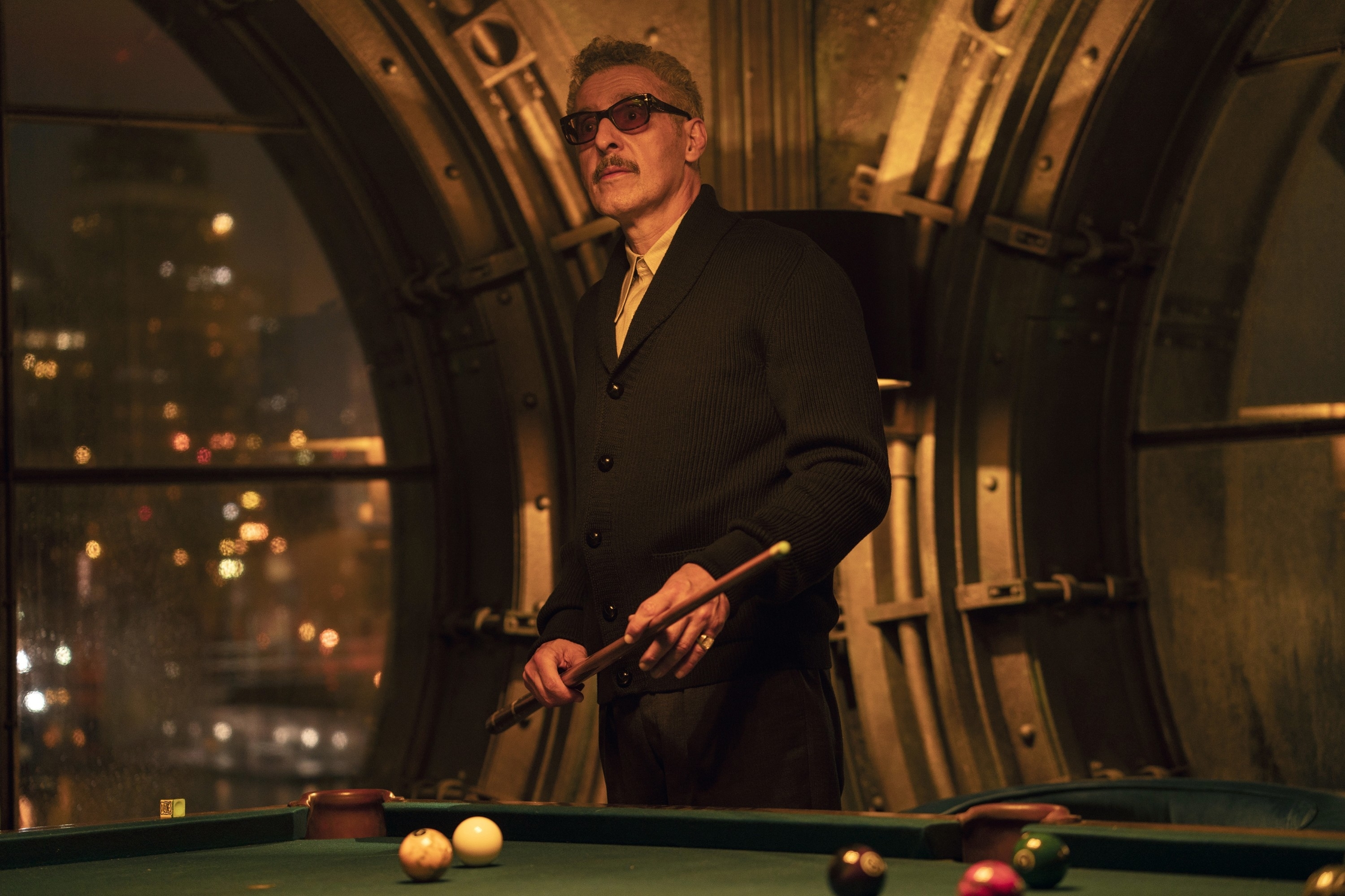 Carmine standing by a pool table in a dimly lit room with a cityscape visible through the round window behind