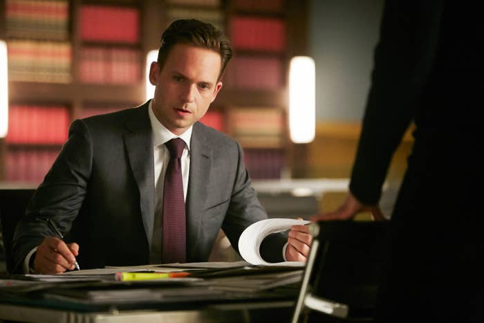 Suit-clad man in an office leans over a desk with papers, talking to an unseen person