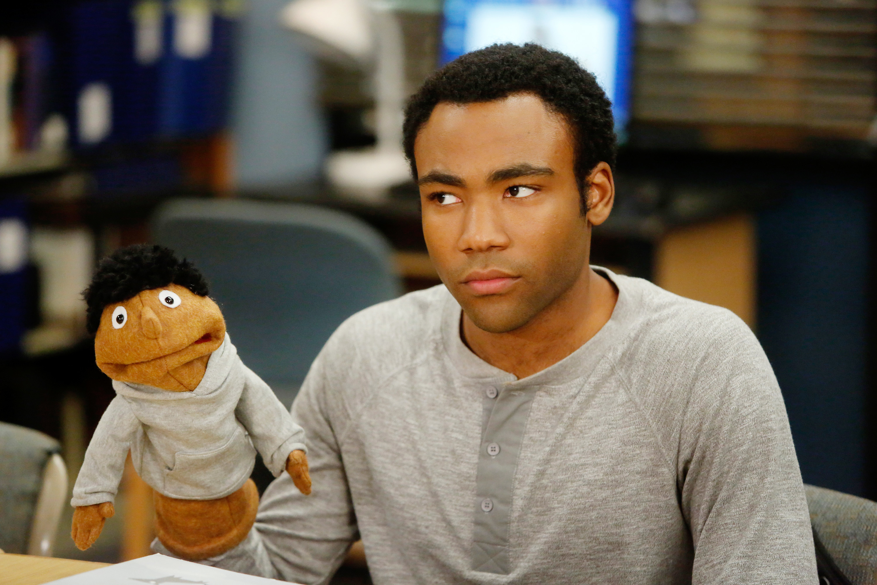 Actor with puppet resembling him on TV show set. He wears a casual grey shirt and looks thoughtful