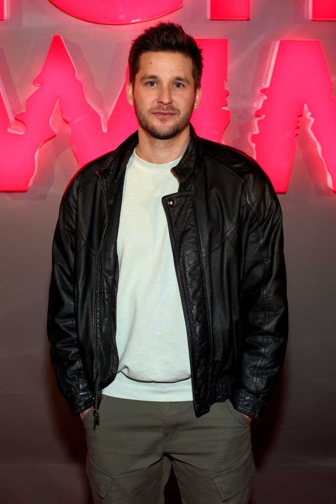 Devon in a black leather jacket over a white t-shirt posing in front of a lit sign
