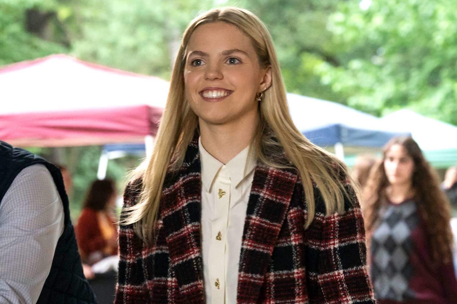 Actress in a scene, wearing a plaid blazer and a smile, with extras in background