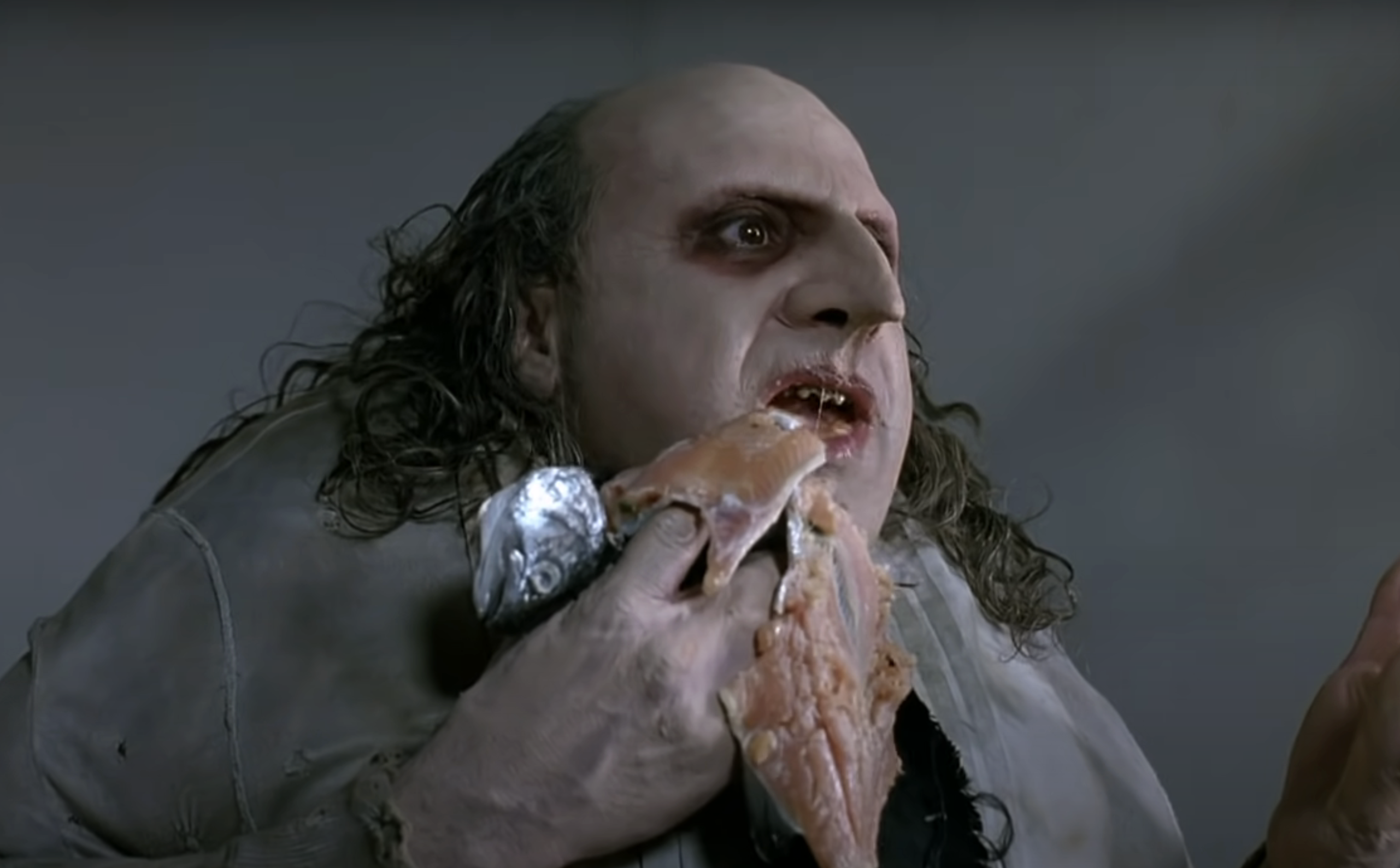 Character portrayed by Danny DeVito in &quot;Batman Returns&quot; grips a fish with his mouth, wearing a stained shirt and coat with frilled edges