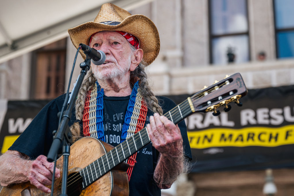 Willie Nelson in a Western-style hat plays guitar and sings into a microphone onstage