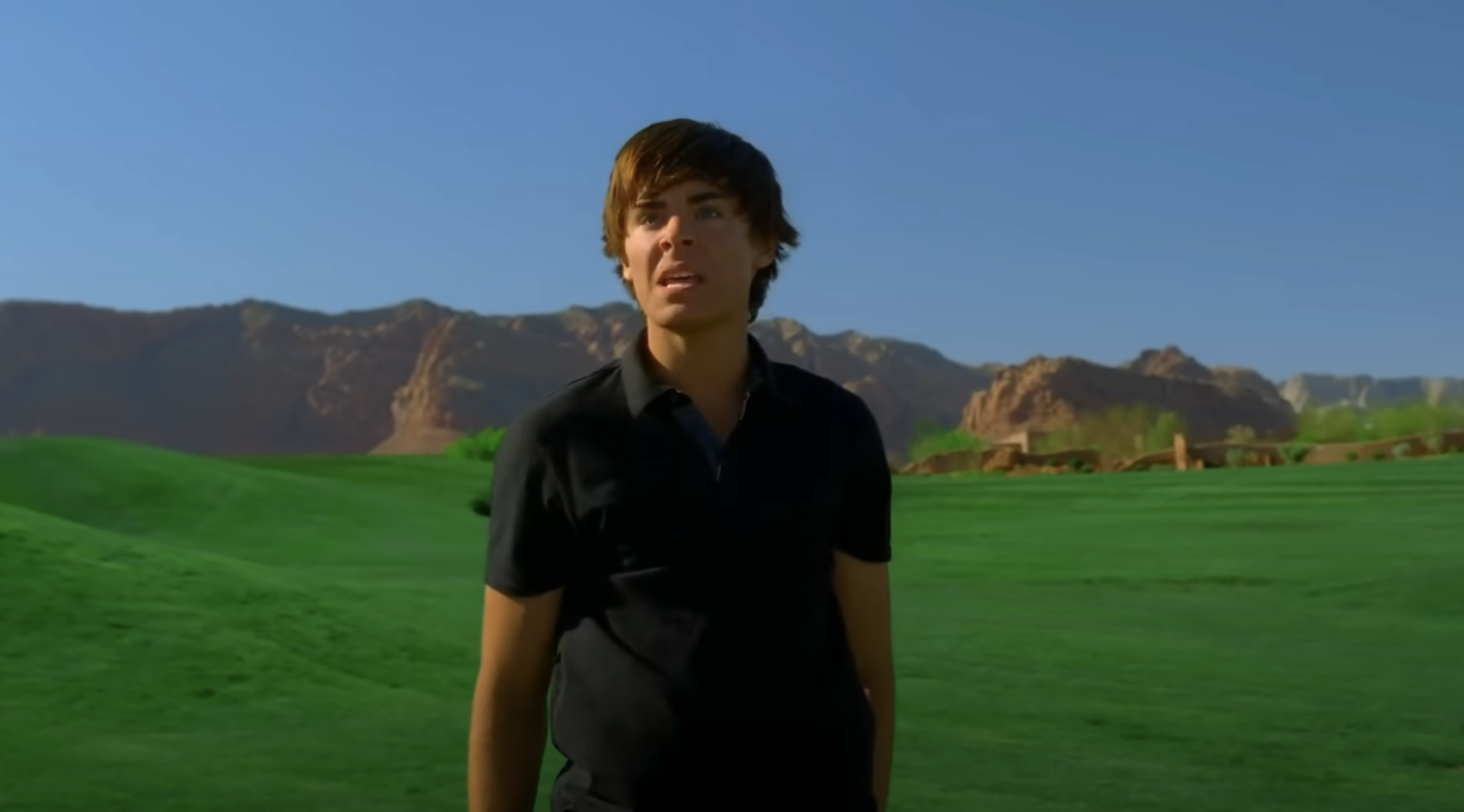 Zac Efron on a golf course with confusion on his face, mountains in the background