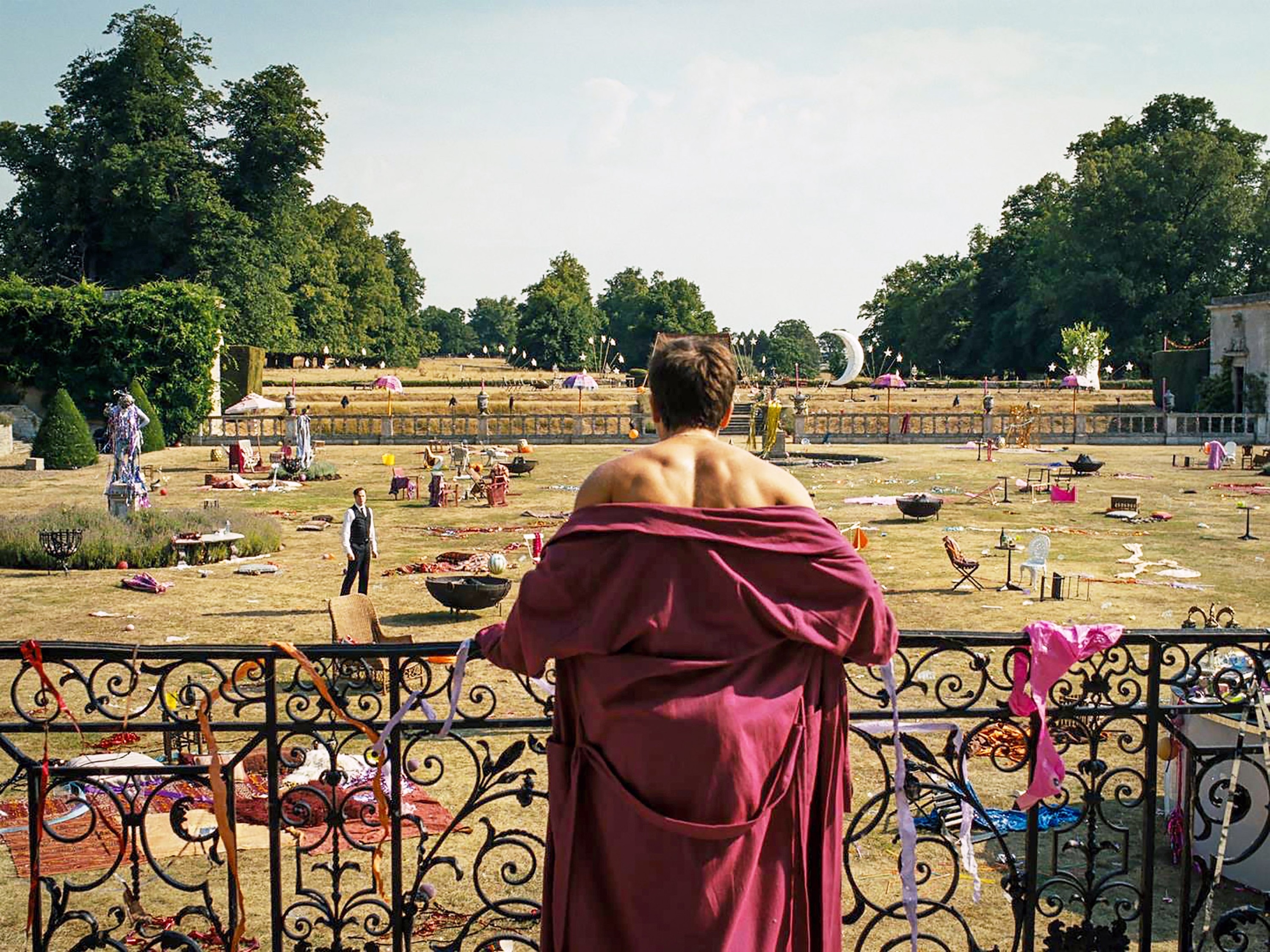 Barry Keogh in burgundy robe overlooking a park with people and scattered objects in Saltburn