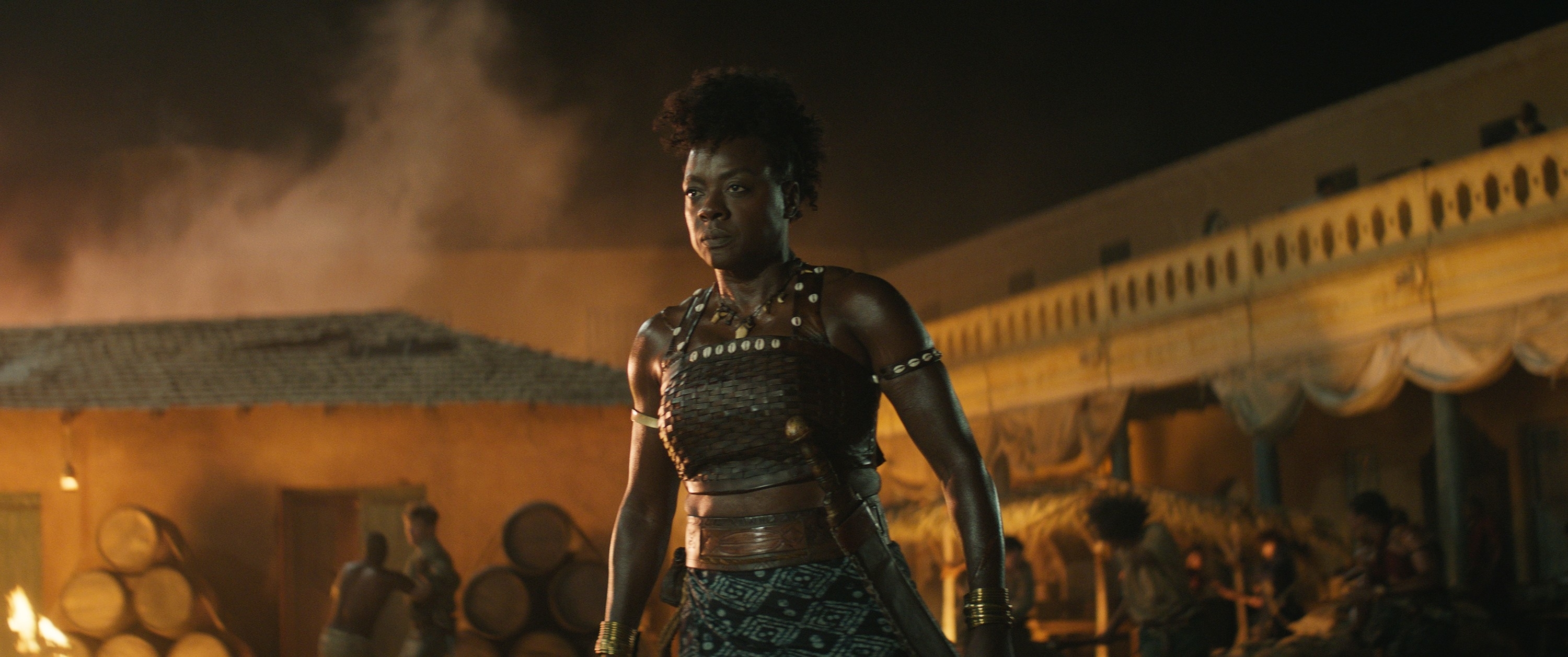Viola Davis in traditional warrior attire stands confidently in a bustling village setting at night