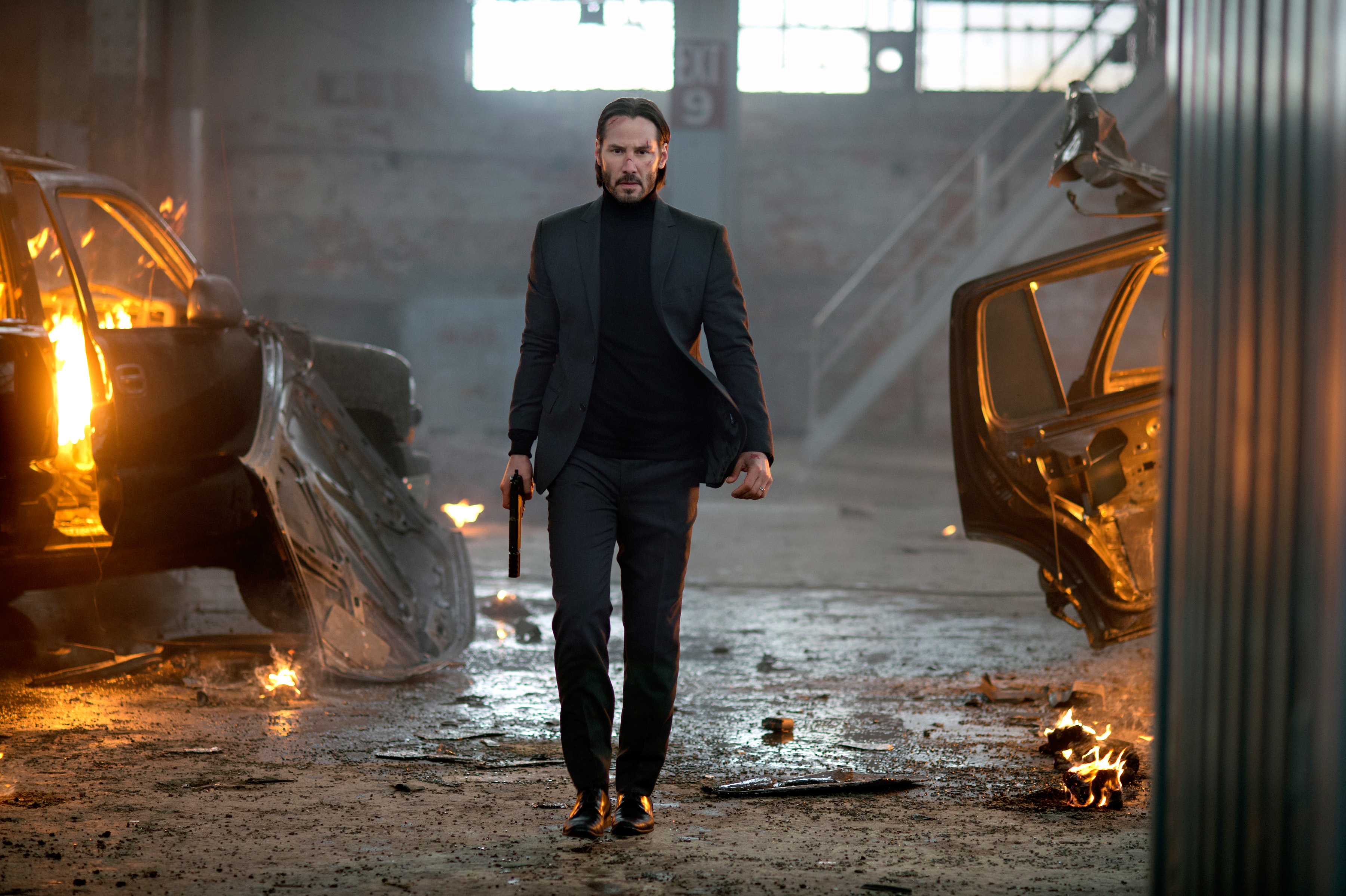 Keanu Reeves as John Wick walking determinedly in a dimly lit space with damaged cars and debris around