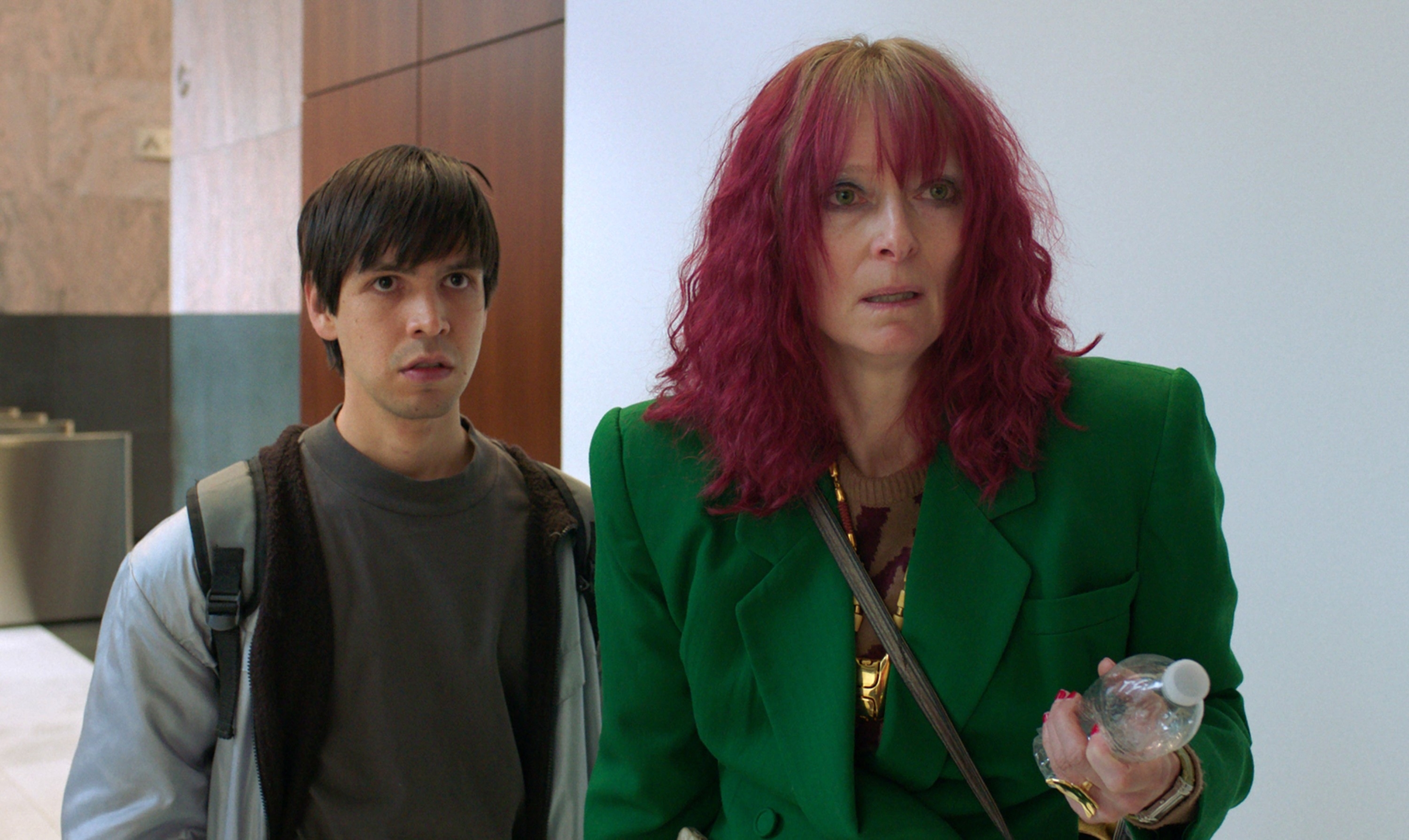 Two characters in a scene, woman in green blazer with pink hair, man in brown shirt with backpack