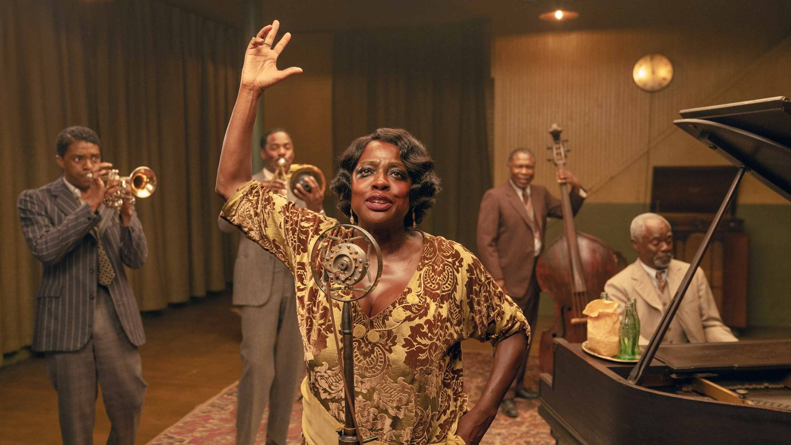 Viola Davis as Ma Rainey in patterned dress singing with jazz band members playing instruments in vintage room