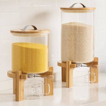 Two clear food storage dispensers with wooden lids and stands, containing grains, with measuring cups below