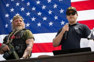 Two individuals stand before an American flag; one speaks into a microphone while the other, with a beard and tattoos, carries a rifle