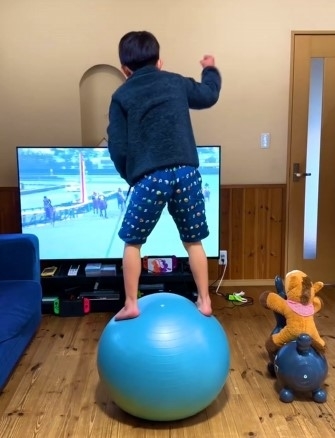 男の子が大きな青いボールの上に立ち、テレビを見ている。