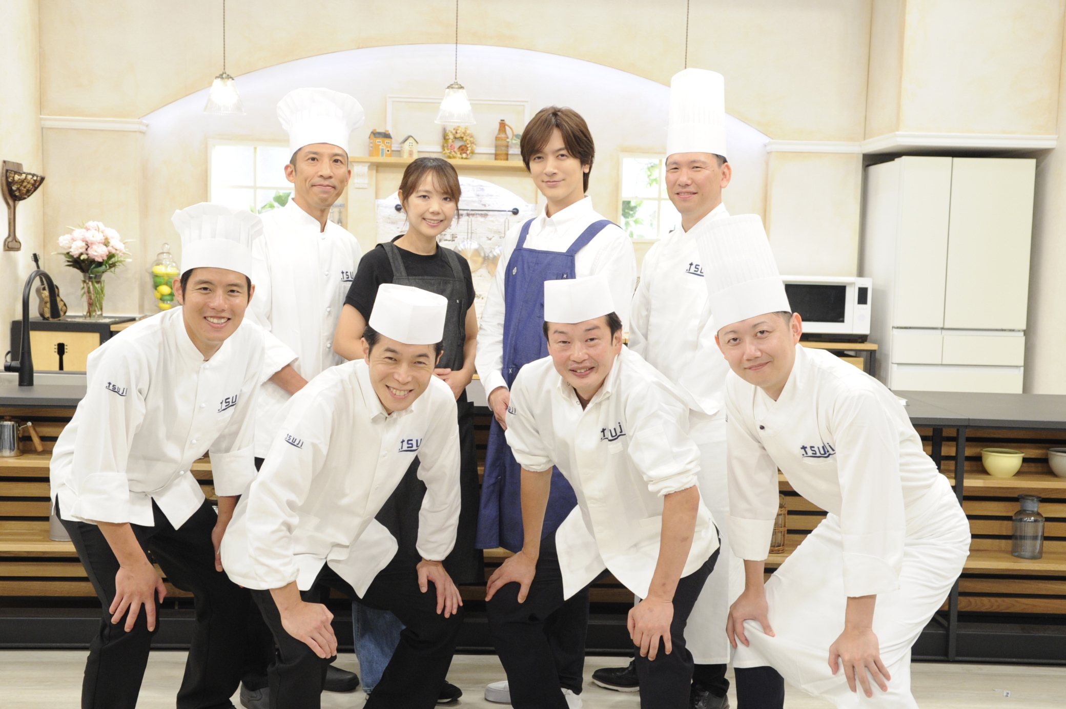 写真はキッチンセットの前で笑顔で立つ8人のシェフ。中央に背の高い男性が目立つ。