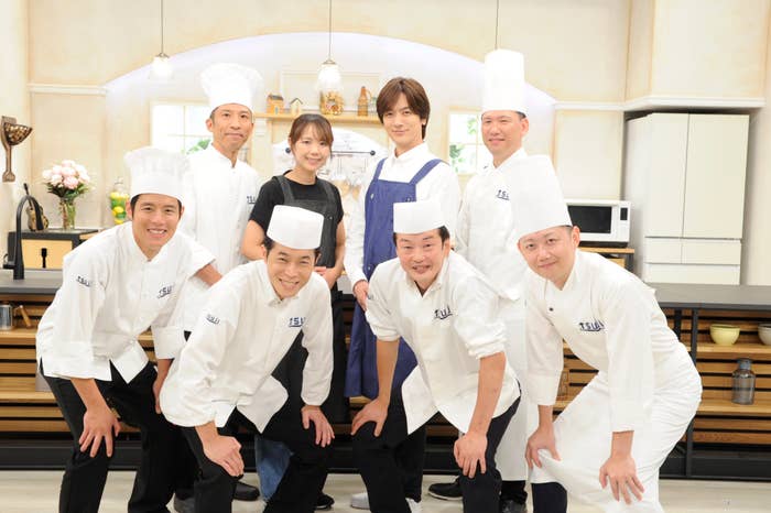 写真はキッチンセットの前で笑顔で立つ8人のシェフ。中央に背の高い男性が目立つ。