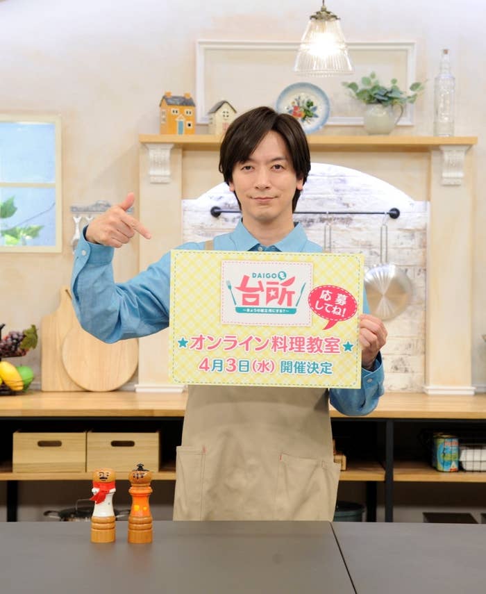 男性がエプロンを着て、番組の宣伝ボードを持ってポーズをとっています。