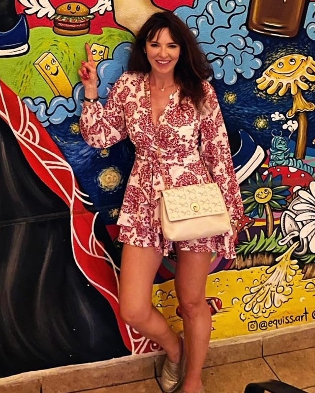 reviewer in a patterned dress posing with a handbag, standing in front of a mural with various painted objects