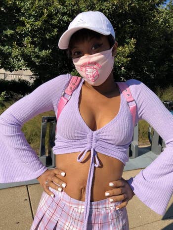 a reviewer outdoors in a lilac crop top and plaid pants, wearing a cap and patterned face mask. Hands on hips