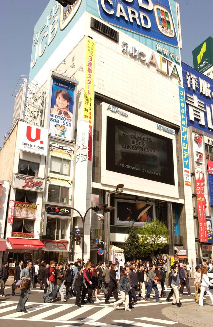 交差点を渡る多くの歩行者と、看板やデジタル広告が見える都会の景色です。