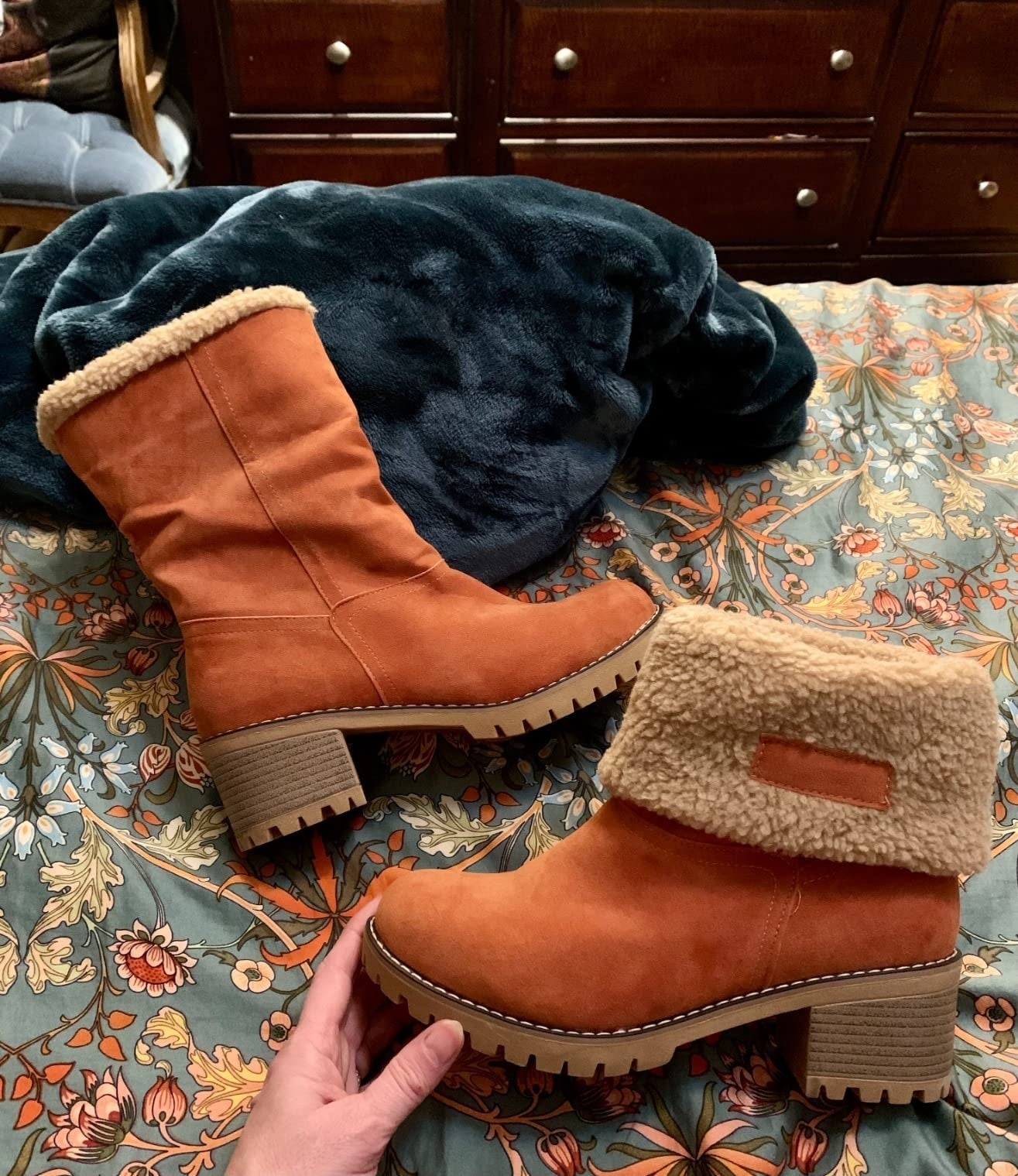 A pair of boots with fuzzy lining and chunky soles rests on a floral bedspread, one standing and one fallen to the side