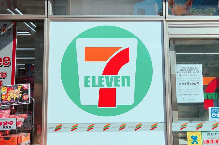 セブンイレブンのロゴが中央にあり、店舗の窓に掲示されています。