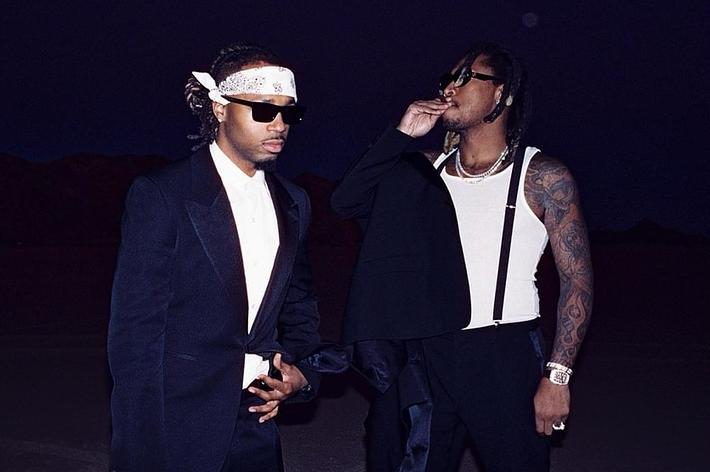 Two men in stylish suits stand confidently, one with sunglasses and a headscarf, the other with visible tattoos